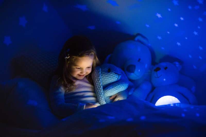Little girl reading a book in bed. Dark bedroom with night light projecting stars on room ceiling. Kids nursery and bedding. Children read before bedtime. Toddler child playing with lamp and bear toy.