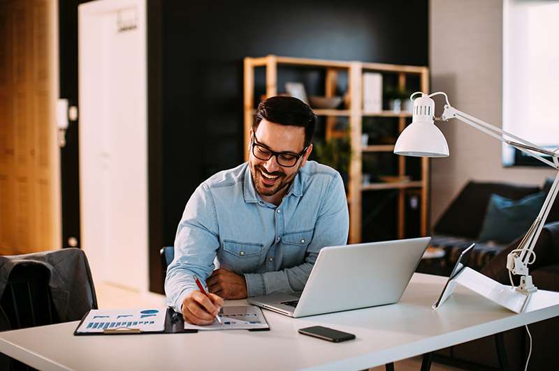 Man writing in notebook looking over files on video call.